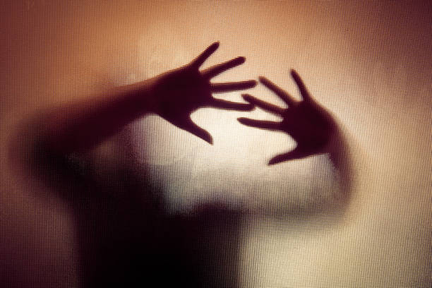 Colour backlit image of the silhouette of a woman with her hands pressed against a glass window. The silhouette is distorted, and the arms elongated, giving an alien-like quality. The image is sinister and foreboding, with an element of horror. It is as if the 'woman' is trying to escape from behind the glass. Horizontal image with copy space.