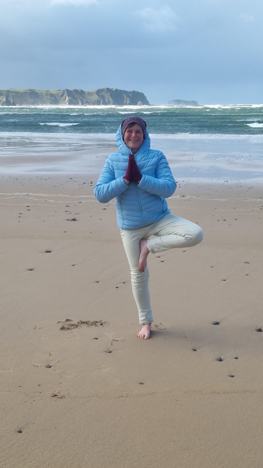 Janet standing in Tree pose, a one legged yoga balance pose on a beach. She is wearing a blue jacket and hat and tousers it appears very windy with big waves. The sky is blue.