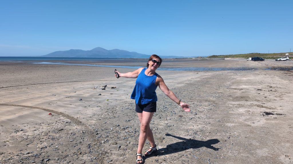woman on the beach with arms extended, blue sky and mountainous background