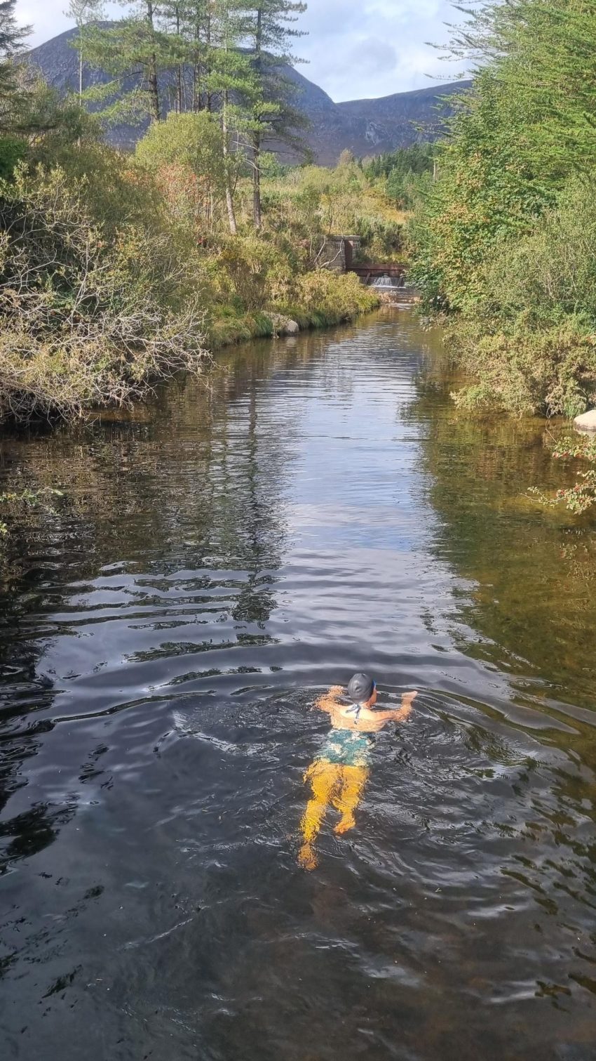 woman swimming up a river surrounded by trees and bushes otherside. The water has a slightly yellow tinge.